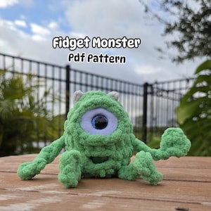May include: A green crocheted monster toy with one large blue eye. The toy is sitting on a wooden surface. The text "Fidget Monster Pdf Pattern" is above the toy.