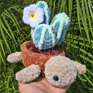 May include: A crocheted turtle holding a blue and green cactus with a light blue flower on top. The turtle is brown and gray and has a black eye.