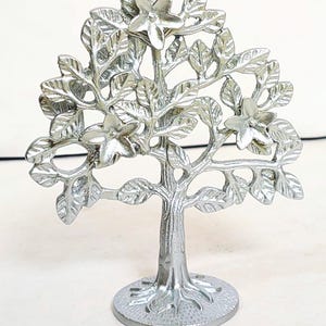 Può includere: Una scultura ad albero in metallo color argento con foglie e fiori dettagliati. L'albero ha una base rotonda e un tronco strutturato. Le foglie e i fiori sono finemente progettati, creando un pezzo decorativo.