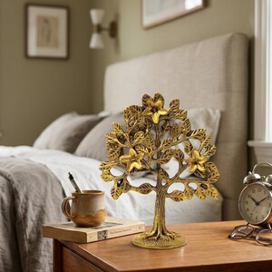 Op de afbeelding: Een goudkleurige metalen boomsculptuur met ingewikkelde details van bladeren en bloemen. De decoratieve boom staat op een houten nachtkastje, naast een mok, boek en wekker. De boom is ongeveer 25 cm hoog.