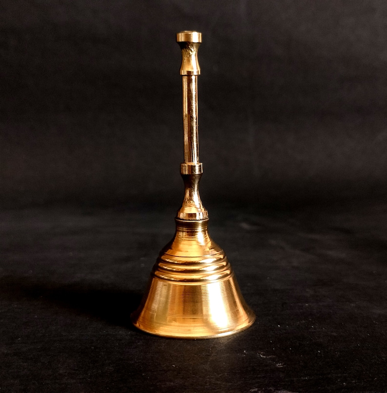 Puede incluir: Una campana de metal de color dorado con una base redonda y un mango largo y delgado.