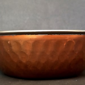 May include: A copper serving bowl with two brass handles. The bowl has a hammered texture and a silver rim.