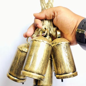 Puede incluir: Un conjunto de cinco campanas de metal de color dorado atadas con una cuerda de yute natural. Las campanas son de forma cilíndrica y tienen un acabado ligeramente desgastado.
