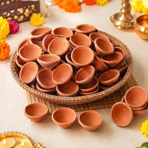 May include: A large collection of small, round, terracotta clay oil lamps, known as diyas, piled inside a woven basket. Several individual terracotta lamps are arranged in front of the basket on a flat surface, showcasing their natural brown colour.