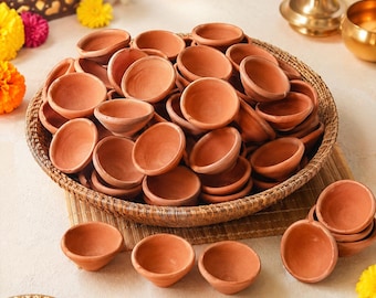 May include: A large collection of small, round, terracotta clay oil lamps, known as diyas, piled inside a woven basket. Several individual terracotta lamps are arranged in front of the basket on a flat surface, showcasing their natural brown color.