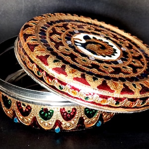 May include: A round, decorative box with a red, gold and green design. The lid is open and the inside of the box is silver. The box is decorated with intricate patterns and jewels.