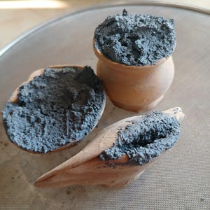 May include: Three small, unglazed terracotta pottery pieces filled with a blue-gray substance. The pieces include a small pot, a halved bowl, and a bird-shaped item, all resting on a metal mesh surface. The pottery has a rustic, handmade appearance.