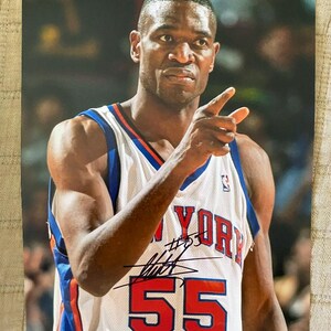 Può includere: Fotografia autografata di un giocatore di basket con una maglia dei New York Knicks. La maglia è bianca con dettagli blu e arancioni, e il numero 55 è ben visibile. Il giocatore indica con il dito indice destro.