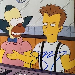 Op de afbeelding: Een geel cartoonfiguur met een rode neus en een blauwe strik staat naast een geel cartoonfiguur met bruin haar. De figuren staan voor een mengpaneel met faders. De afbeelding is gesigneerd met "Matt Groening".