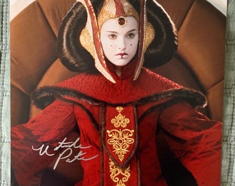 May include: A signed photograph of a woman wearing a red and gold costume with a crown. The woman is sitting on a throne and has a serious expression on her face. The signature is "Natalie Portman".