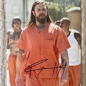 Puede incluir: Una persona con tatuajes y barba lleva un uniforme naranja de manga corta con un bolsillo. La persona está de pie frente a un fondo borroso con otras personas. La imagen tiene una firma en tinta negra.