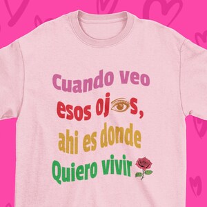 May include: Pink t-shirt with a Spanish phrase that reads "Cuando veo esos ojos, ahi es donde quiero vivir" which translates to "When I see those eyes, that's where I want to live". The phrase is in colorful text with a small red rose graphic.