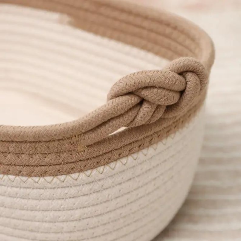 May include: Close-up of a woven rope basket with a knotted handle. The basket features a two-tone design, with a cream-colored interior and a tan-colored rope trim and handle. The basket is made of a soft, textured material.
