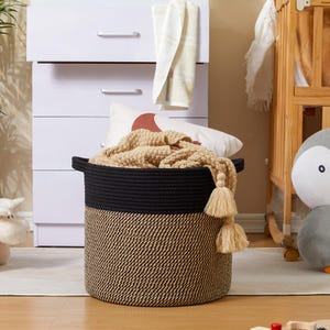 Peut inclure: Un panier de rangement rond avec un dessus noir et un fond tissé beige et noir. Le panier est rempli d'une couverture beige et comporte deux glands beiges. Une commode blanche et un berceau sont en arrière-plan.