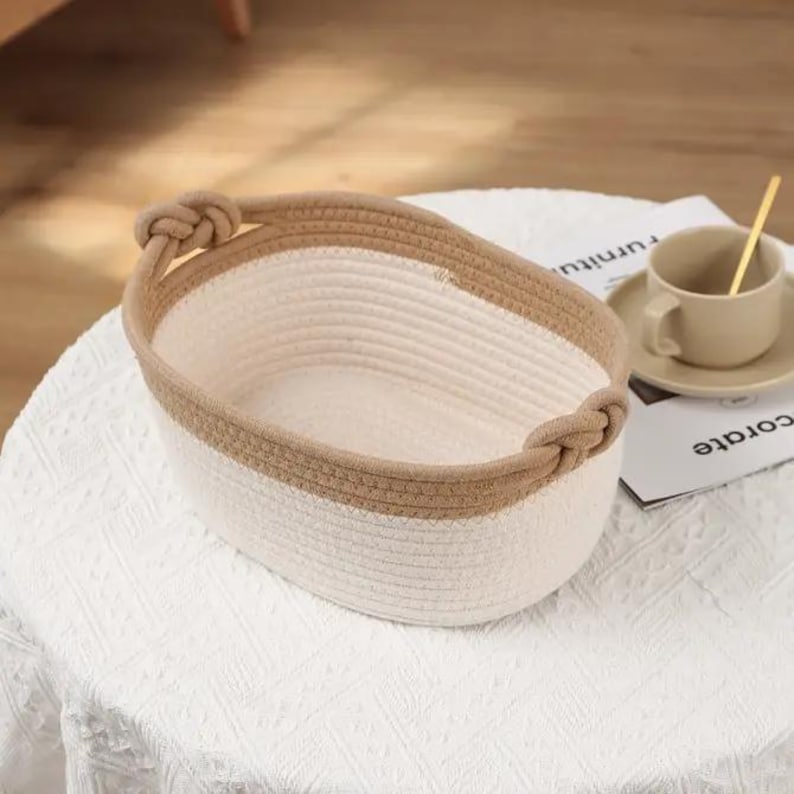 May include: Oval-shaped woven basket with a white interior and a tan border. The basket has two knotted handles on either side. The basket is sitting on a white, textured surface.