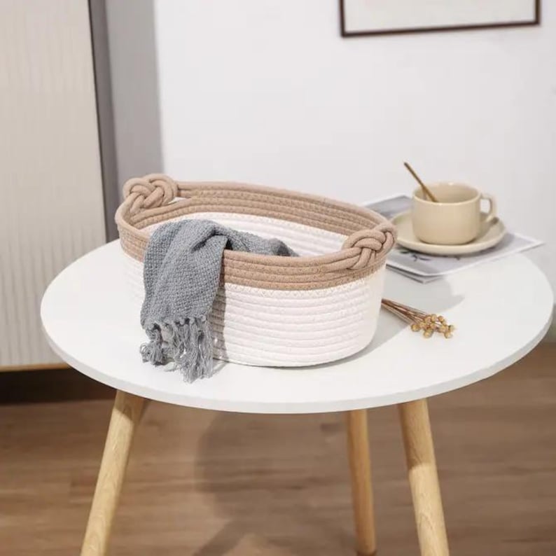 May include: A white and tan oval-shaped woven basket with knotted handles. A gray fringed scarf is inside the basket. The basket sits on a white table with wooden legs. A cup and saucer are on the table.