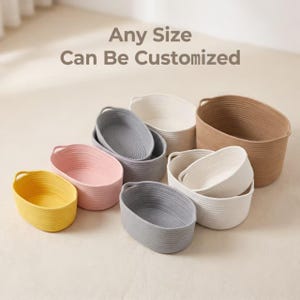 Peut inclure: Une collection de paniers de rangement ovales en corde torsadée de couleur jaune, rose, gris, blanc et beige. Chaque panier possède deux poignées intégrées. Le texte "Any Size Can Be Customized" est affiché au-dessus des paniers.
