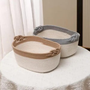 May include: Two oval-shaped rope baskets. One basket has a tan rim and handles, the other has a gray rim and handles. Both baskets have a white interior and are sitting on a white surface.