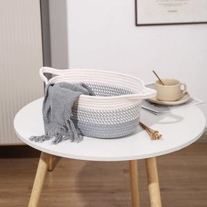May include: A round, woven basket with a white and gray color scheme, featuring two handles. A gray blanket with fringe is draped over the side. The basket sits on a white table with wooden legs, alongside a teacup and saucer.