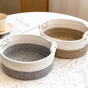 Puede incluir: Dos cestas redondas de cuerda trenzada con asas trenzadas. Una cesta es gris y blanca, la otra marrón y blanca. Las cestas están sobre un mantel blanco de encaje. Una taza y un platillo están en el fondo.