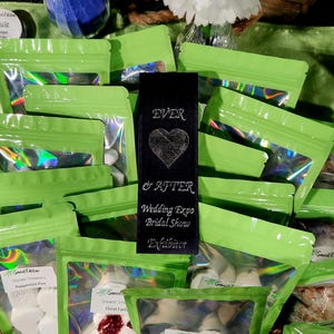May include: Green and clear plastic bags filled with white and pink shower steamers. The bags are labeled with the text "Sweet Edition" and the scent of the steamers. A black bookmark with the text "EVER & AFTER Wedding Expo Bridal Show Exhibitor" is in the center of the image.