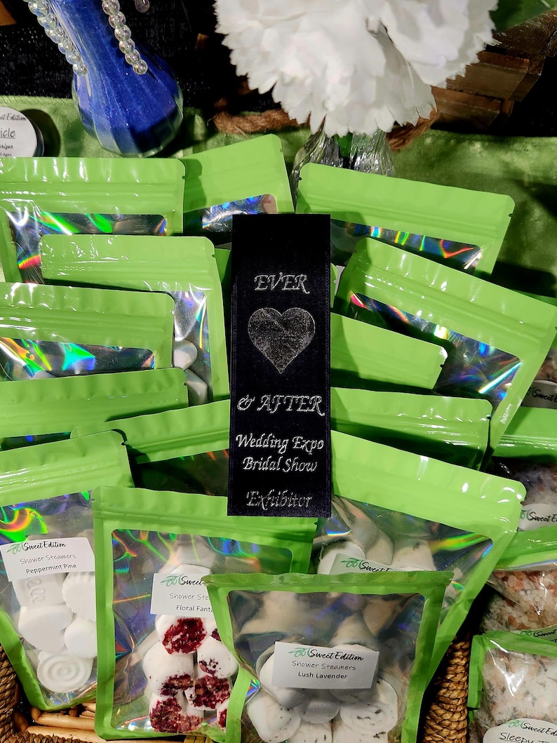 May include: Green and clear plastic bags filled with white and pink shower steamers. The bags are labeled with the text "Sweet Edition" and the scent of the steamers. A black bookmark with the text "EVER & AFTER Wedding Expo Bridal Show Exhibitor" is in the center of the image.