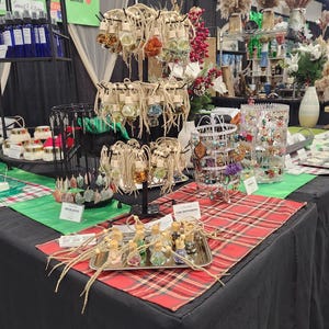 May include: A table display of handmade jewelry and car air fresheners. The jewelry includes necklaces with colorful beads and charms. The car air fresheners are in small glass bottles with cork tops and colorful crystals inside. The table is covered with a red and black plaid tablecloth.