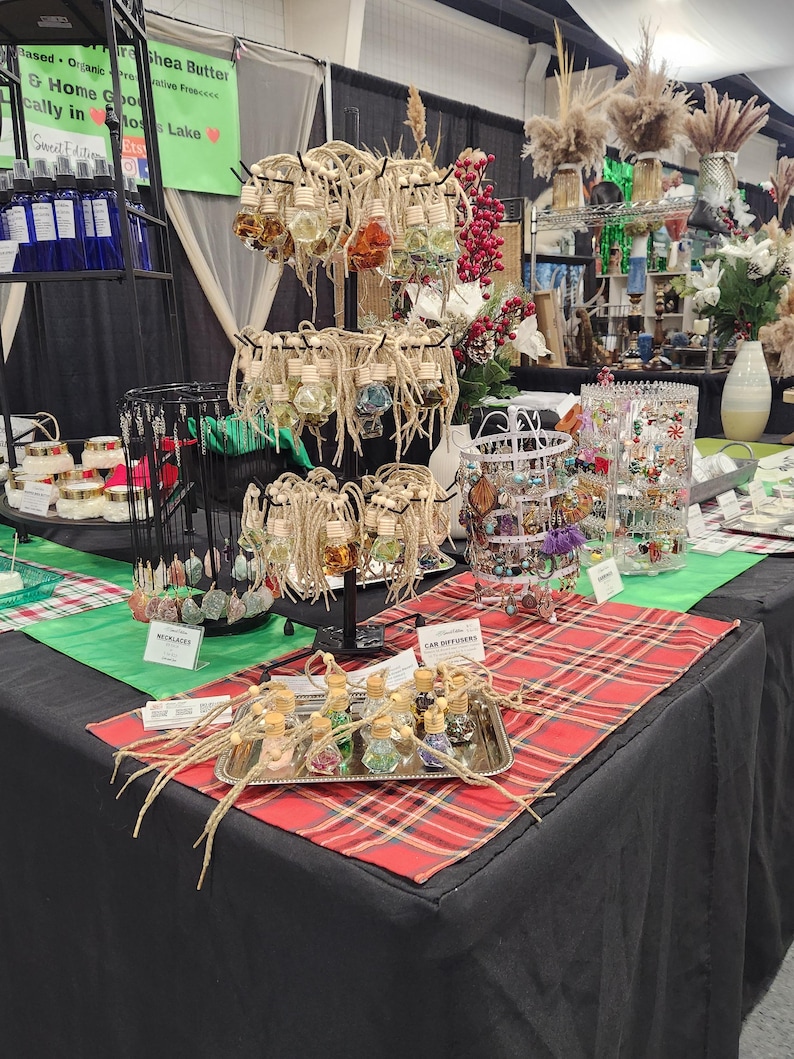 May include: A table display of handmade jewelry and car air fresheners. The jewelry includes necklaces with colorful beads and charms. The car air fresheners are in small glass bottles with cork tops and colorful crystals inside. The table is covered with a red and black plaid tablecloth.