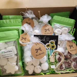 May include: A gift box filled with handmade shower steamers and a car air freshener. The shower steamers are in various shapes and sizes, including stars, hearts, and circles. The car air freshener is a small glass bottle with a wooden cap and a string attached. The box is lined with green shredded paper and has a "Thank You" card with sunflowers on it.