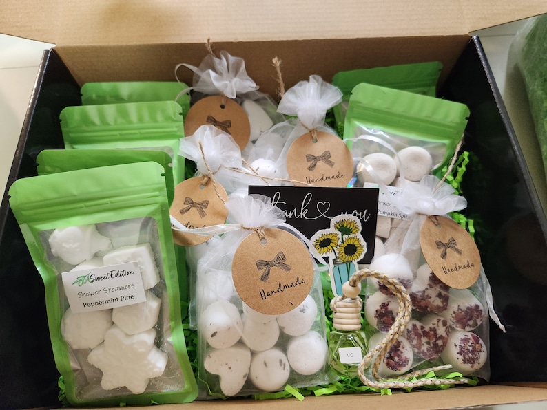 May include: A gift box filled with handmade shower steamers and a car air freshener. The shower steamers are in various shapes and sizes, including stars, hearts, and circles. The car air freshener is a small glass bottle with a wooden cap and a string attached. The box is lined with green shredded paper and has a "Thank You" card with sunflowers on it.