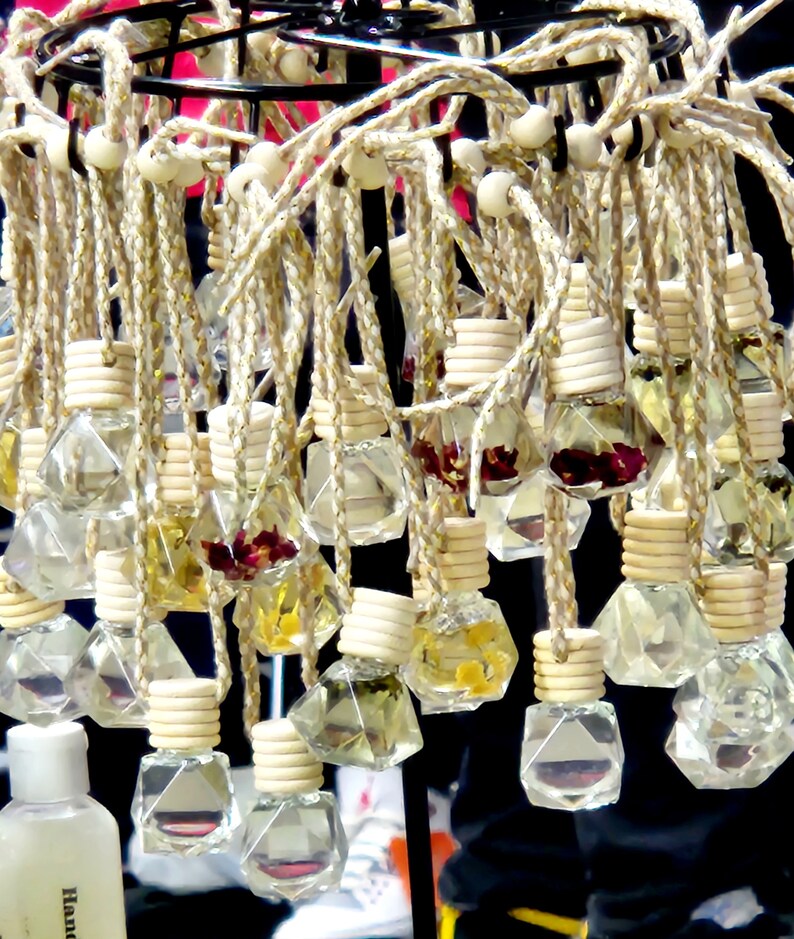 May include: A display of small glass bottles with various dried flowers and herbs inside, hanging from a metal rack with white cords. The bottles are clear and have a diamond shape.