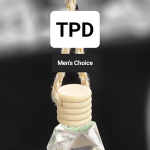 May include: A clear glass bottle with a geometric shape hangs from a beige rope with gold accents. The bottle is attached to a wooden disc with a beige rope. The bottle has a label that says "TPD Men's Choice".