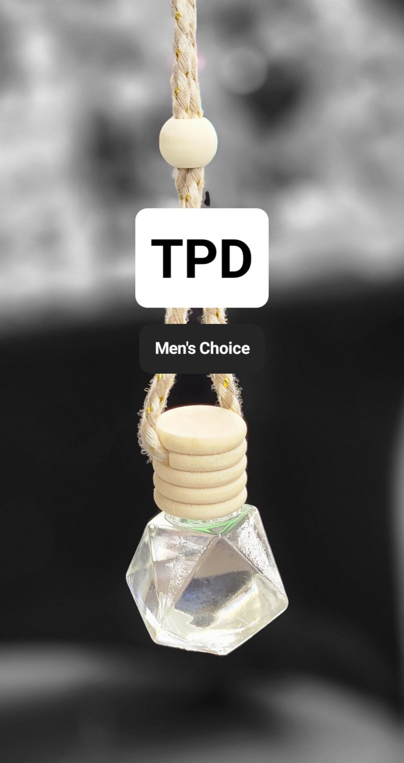 May include: A clear glass bottle with a geometric shape hangs from a beige rope with gold accents. The bottle is attached to a wooden disc with a beige rope. The bottle has a label that says "TPD Men's Choice".