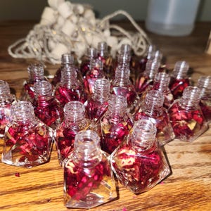 May include: A collection of small, clear glass bottles filled with dried red rose petals and a clear liquid. The bottles are shaped like diamonds and have a cork stopper.