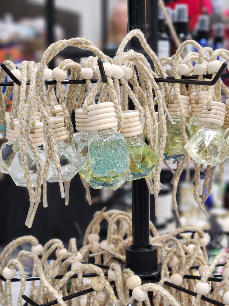 May include: A display of decorative car air fresheners with a variety of colors and designs. The air fresheners are made of glass and have a geometric shape. They are hanging from a metal rack with twine and wooden beads.