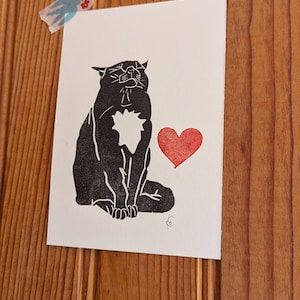 Puede incluir: Un grabado en linóleo de un gato negro con un corazón rojo. El gato está sentado con los ojos cerrados y un recorte en forma de corazón en el pecho. La impresión está sobre papel blanco y pegada a una superficie de madera.