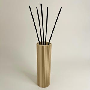 May include: A beige reed diffuser with black reeds. The cylindrical diffuser has vertical ribbed detailing. The reeds are black and extend upwards from the diffuser. The background is a solid white.