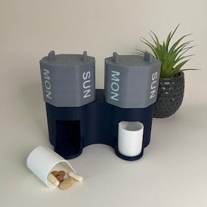 Könnte beinhalten: Ein zweiteiliger Pillenorganizer in Marineblau und Grau. Jeder Abschnitt ist mit "MON" und "SUN" in Weiß beschriftet. Ein kleiner weißer Becher und ein verschütteter Behälter mit Pillen befinden sich im Vordergrund. Eine kleine Topfpflanze ist im Hintergrund.