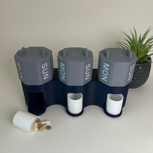 Puede incluir: Un organizador de pastillas de tres secciones en gris y azul marino. Cada sección está etiquetada con "LUN" y "DOM" y tiene una tapa con un icono de sol, luna o nube. Dos vasos blancos están en la base, y un tercer vaso está volcado, derramando pastillas.