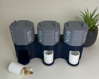 May include: A three-section pill organizer in gray and navy blue. Each section is labeled with "MON" and "SUN" and has a lid with a sun, moon, or cloud icon. Two white cups are in the base, and a third cup is tipped over, spilling pills.