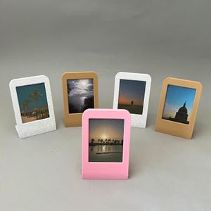 May include: Five small, rectangular, upright picture frames in various colors including white, tan, and pink. Each frame holds a different landscape photograph, featuring scenes such as a beach with palm trees, a sunset, and a building silhouette.