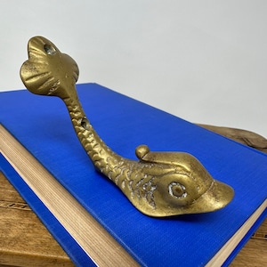 May include: A brass fish-shaped hook with a detailed design. The hook is on a blue book.