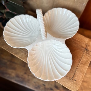 Może przedstawiać: Biała ceramiczna tacka do serwowania w kształcie trzech muszli z plecionym uchwytem z sznurka.