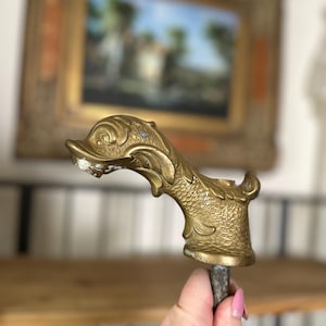 May include: A gold-toned metal faucet spout shaped like a fish with detailed scales and fins. The spout is attached to a metal pipe.