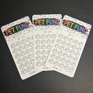 May include: Three white Pet Fund cards with a rainbow-colored "PET FUND" banner. Each card features paw prints and the text "£5", "£10", and "£". The cards are designed to help save money for pets.