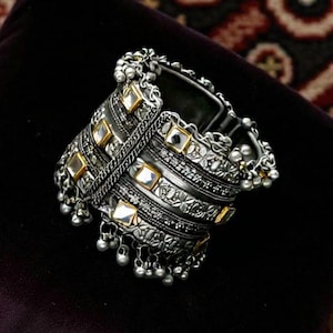 Op de afbeelding: Zilverkleurige manchetarmband met vierkante goudkleurige accenten en bungelende zilveren kralen. De armband heeft ingewikkelde geëtste ontwerpen en wordt weergegeven op een donkerpaars fluwelen oppervlak.