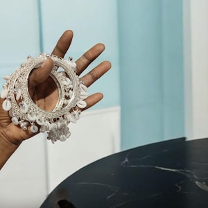 Peut inclure: Bracelets argentés ornés de coquillages blancs sont tenus dans une main. Les bracelets présentent un design circulaire avec des détails complexes et des breloques en coquillages. Le bijou est présenté sur un fond bleu clair.