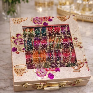 Puede incluir: Una caja decorativa dorada con una ventana transparente, llena de pulseras coloridas. La caja presenta diseños florales y de tazas de té. Las pulseras son de varios colores, incluyendo rojo, verde y naranja.