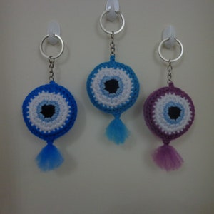 Puede incluir: Tres llaveros de ganchillo con forma de ojo turco, en colores azul, morado y blanco. Cada llavero tiene un borla.