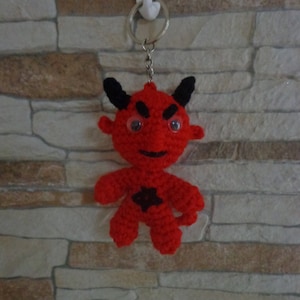 Pode incluir: Chaveiro de crochê de diabo vermelho com chifres pretos, uma estrela preta no peito e olhos saltados.